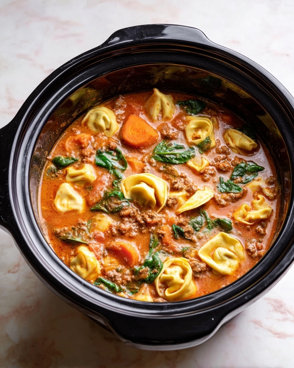 A black slow cooker filled with a rich tomato-based soup showing three main layers: at the top, soft yellow tortellini pasta floating with visible folds and edges, next large slices of orange carrots scattered evenly, and green spinach leaves adding fresh color. The soup’s thick broth is a warm, orange-red with bits of ground meat and vegetable chunks throughout. The cooker is set on a white marbled texture surface, highlighting the colors inside. photo taken with an iphone --ar 4:5 --v 7