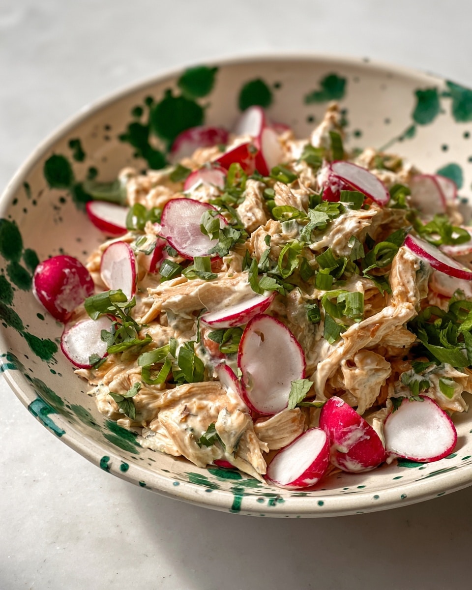 A creamy salad is served in a white bowl with green splatter patterns. The dish has shredded chicken mixed in a thick, off-white creamy sauce, forming the main textured layer. Scattered on top are thinly sliced radishes with bright red edges and white centers, adding fresh pops of color. Small green onion rings and leafy herbs are sprinkled throughout, giving the salad a vibrant green contrast. The bowl rests on a white marbled surface. photo taken with an iphone --ar 4:5 --v 7