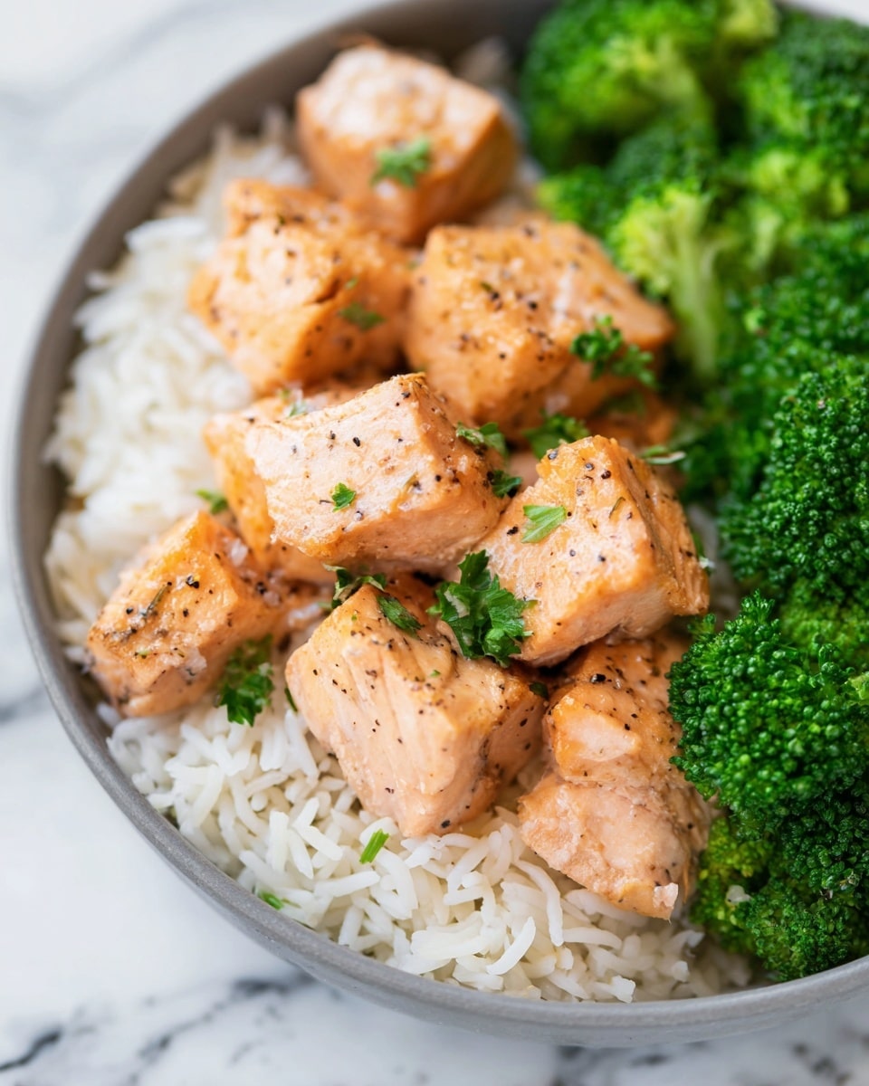 The image shows a dish with three main layers in a white bowl placed on a white marbled surface. The bottom layer is white rice with long grains scattered unevenly. On top of the rice, there are several pieces of cooked salmon cut into bite-sized cubes, with a light orange-pink color and specks of black pepper. The salmon pieces have a slightly firm texture and are topped with small green parsley leaves for garnish. On one side of the bowl, there is a portion of bright green broccoli florets, vibrant and fresh-looking with a bumpy texture. photo taken with an iphone --ar 4:5 --v 7