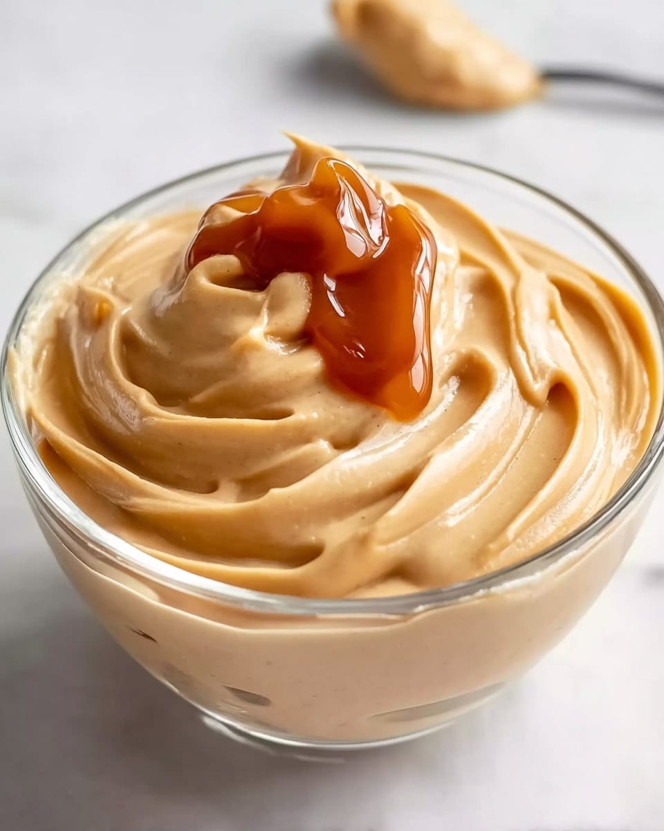 The image shows a clear glass bowl filled with a smooth, creamy light brown spread, swirled gently to create soft peaks and ridges. On top, there is a dollop of shiny, thick amber-colored caramel sauce that contrasts with the matte texture of the spread beneath it. The bowl sits on a white marbled surface, adding subtle texture in the background. The light creates soft shadows, highlighting the swirls and glossy caramel. photo taken with an iphone --ar 4:5 --v 7