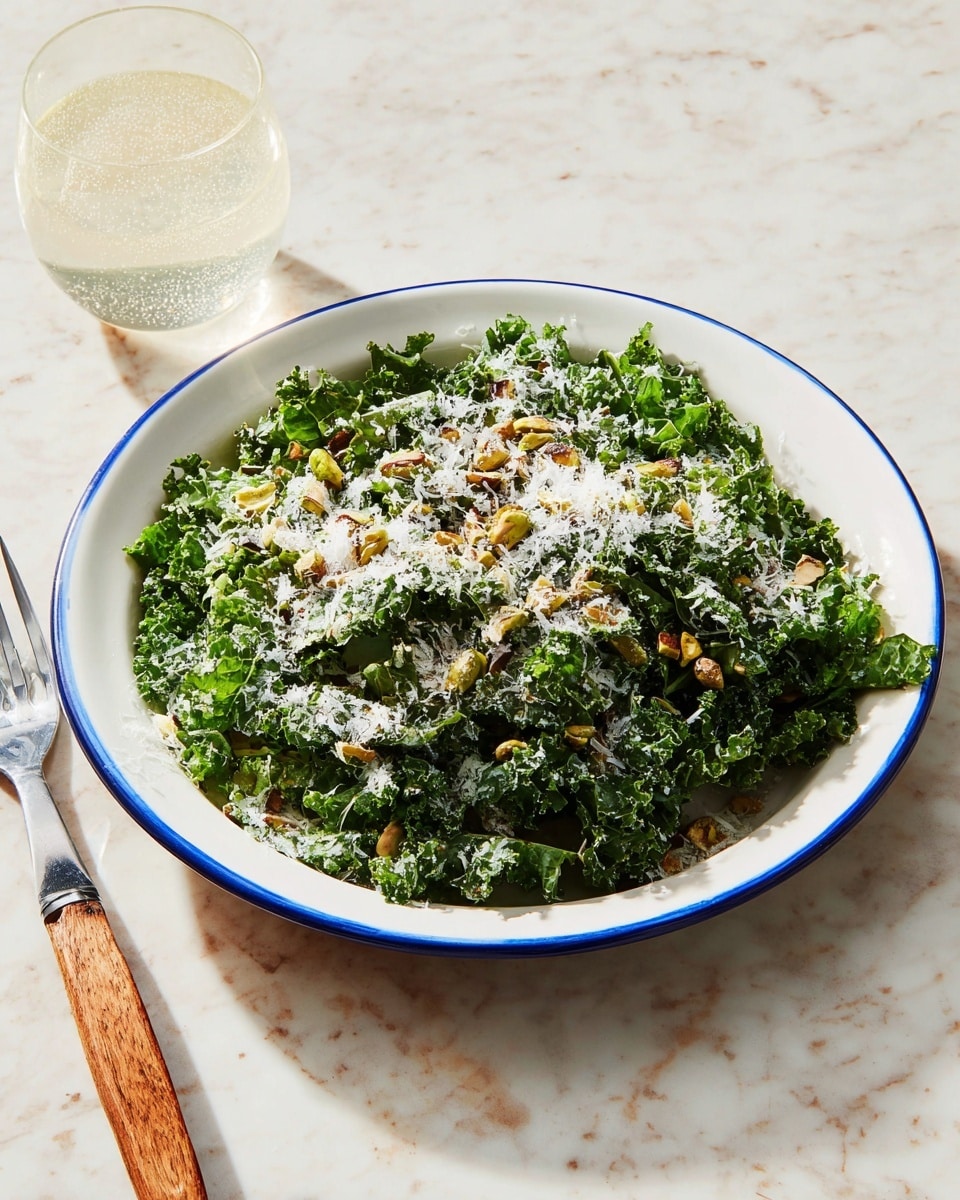 A white plate with a blue rim is filled with a fresh kale salad. The bottom layer is dark green kale leaves, curly and crisp. On top, there are small chopped pistachios scattered, adding a light brown and green texture. Over everything, there is a fine layer of white grated cheese, slightly melting into the kale. The plate is set on a white marbled surface next to a fork with a wooden handle and a glass of sparkling water. The light casts soft shadows. photo taken with an iphone --ar 4:5 --v 7
