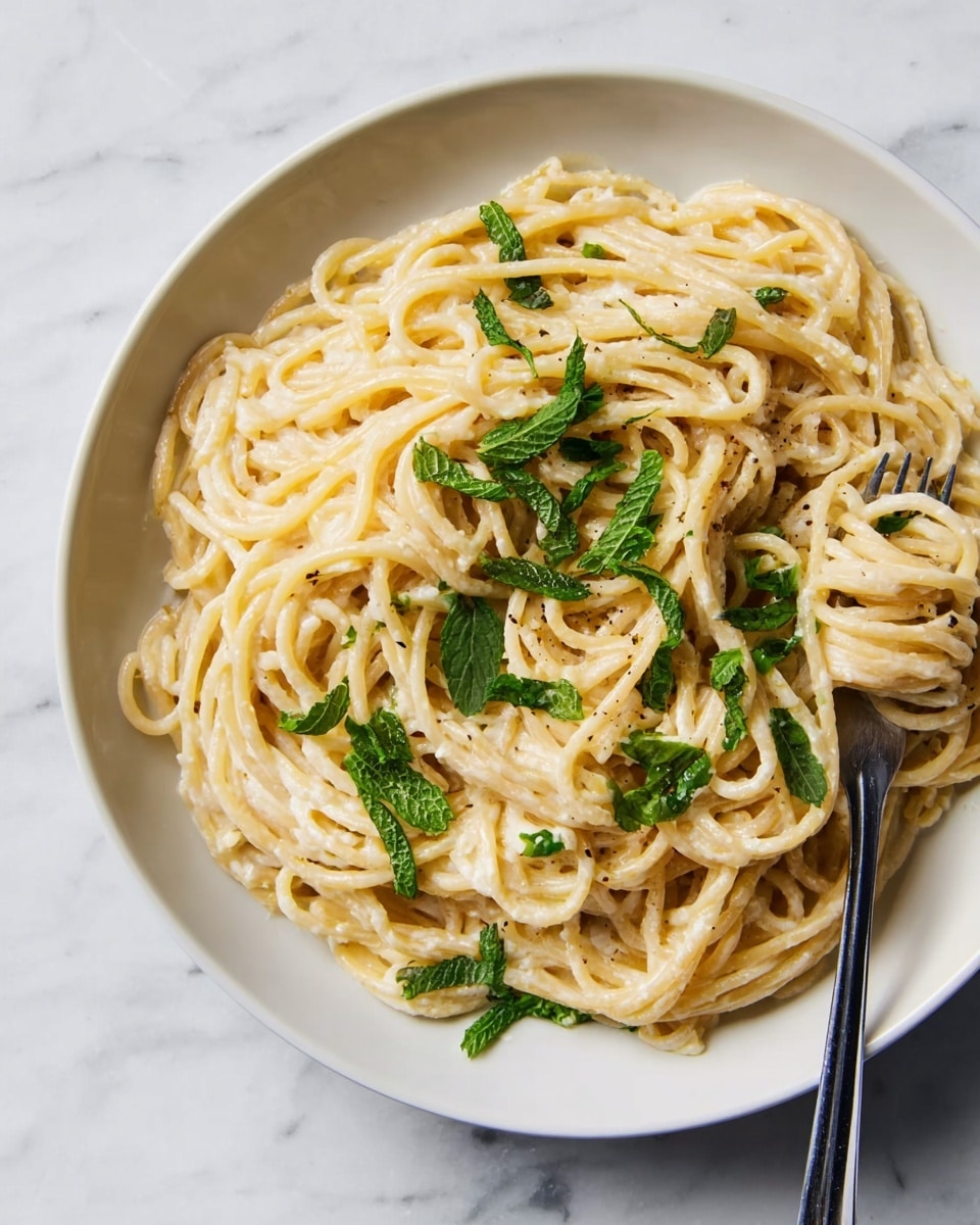 A white shallow bowl contains two layers of thick spaghetti pasta coated with a light cream-colored sauce, giving a smooth and slightly glossy texture. The pasta is intertwined in a loose heap at the center of the bowl. Scattered on top are bright green torn mint leaves providing a fresh pop of color. A silver fork with a black handle rests on the right edge of the bowl, twirling some of the pasta strands. The bowl is set on a white marbled surface that adds a clean and bright backdrop to the dish. Photo taken with an iphone --ar 4:5 --v 7